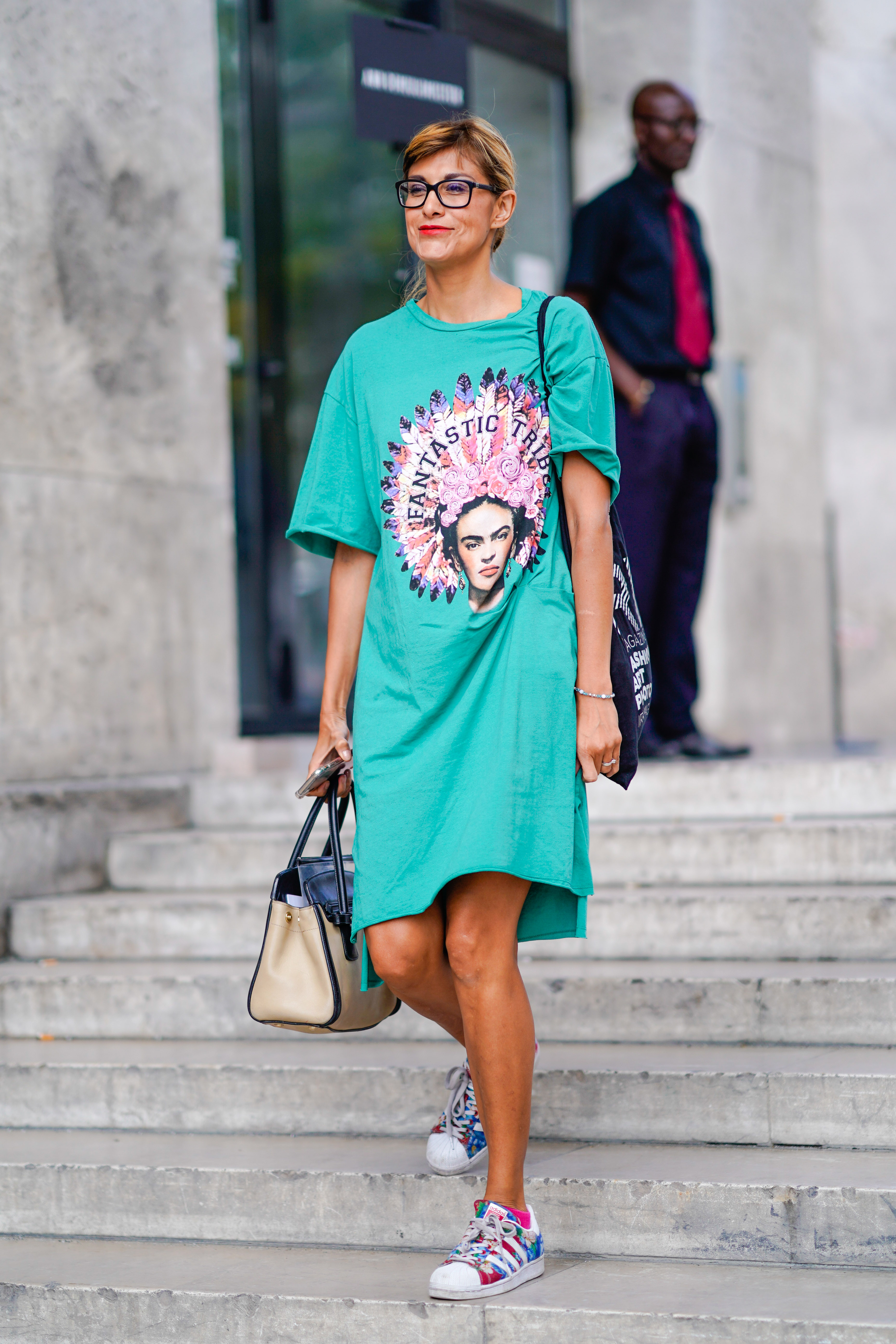 A person in a green t-shirt dress with a Frida Kahlo print, glasses, carrying a beige bag, and wearing colorful sneakers, walks down stairs.