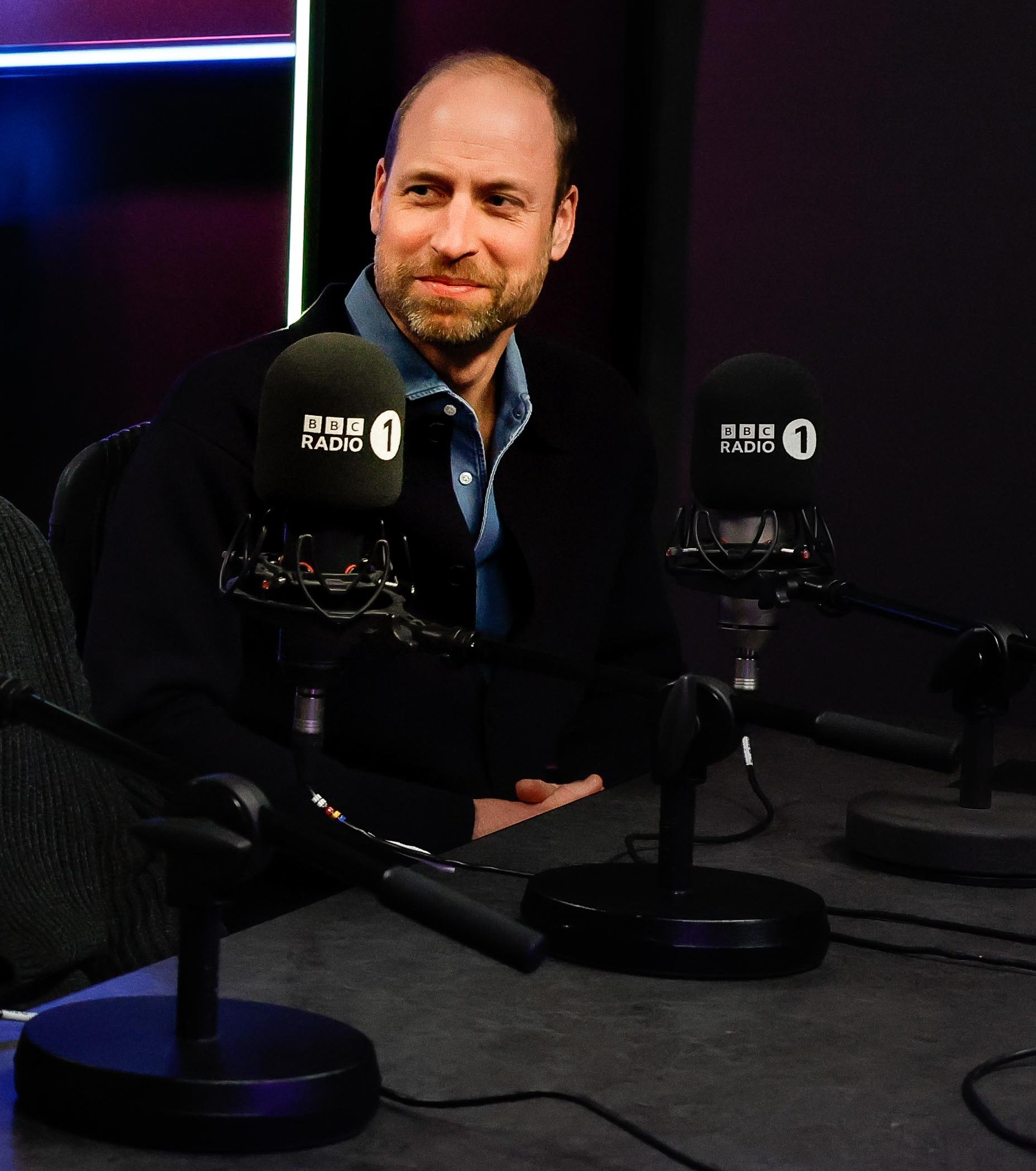 The Prince of Wales smiling and looking to the left in a radio studio with two BBC Radio 1 microphones in front of him.