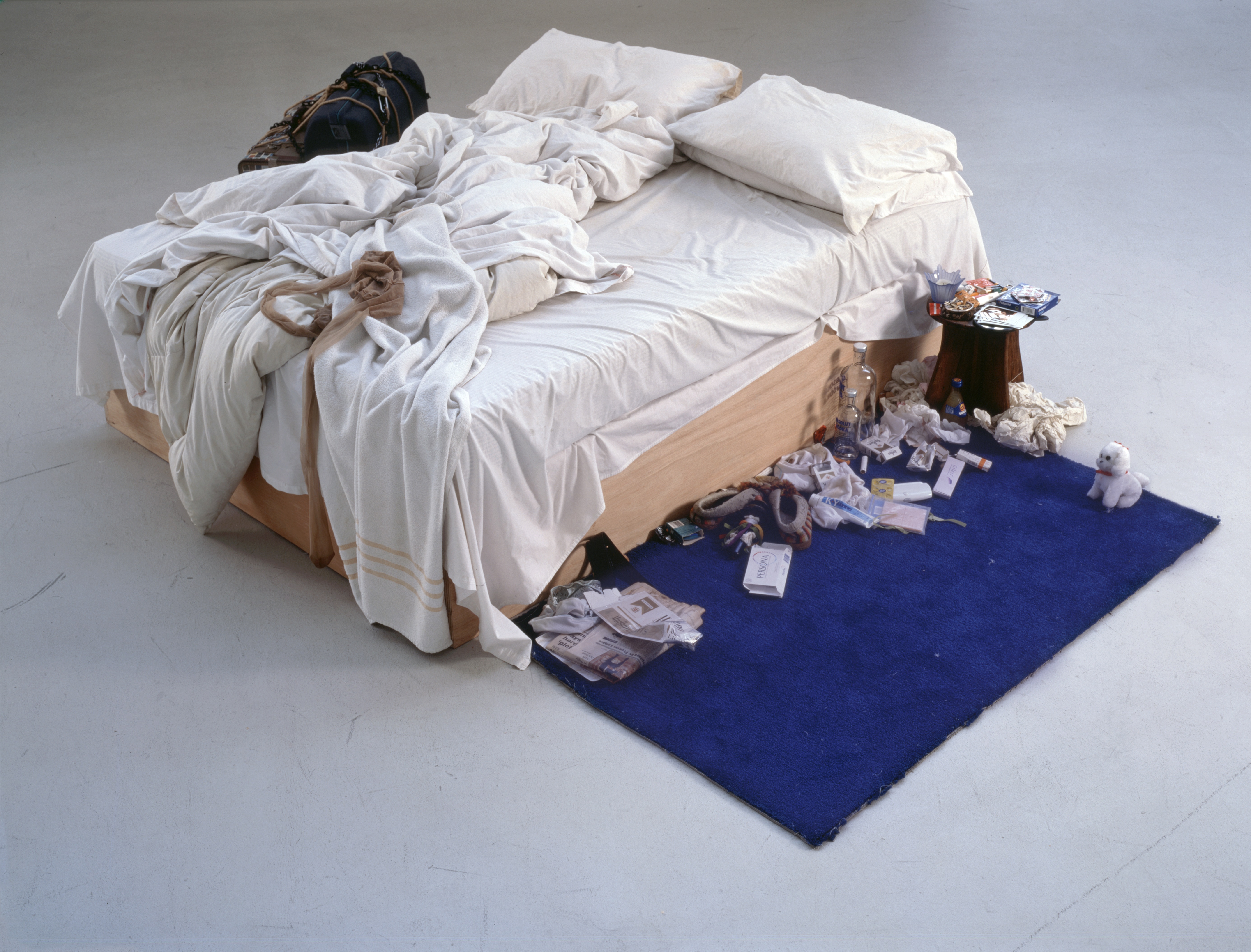 Tracey Emin's "My Bed" art installation, featuring an unmade bed surrounded by discarded personal items and a blue rug.
