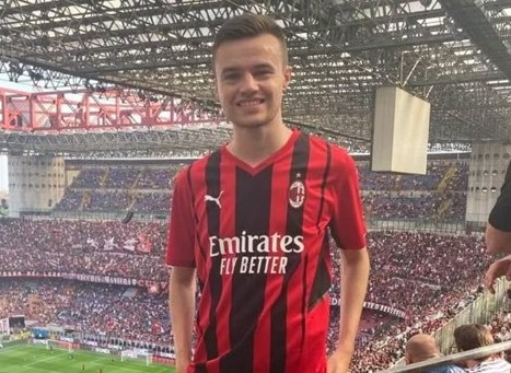 A man in a red and black striped shirt smiling at a football stadium.