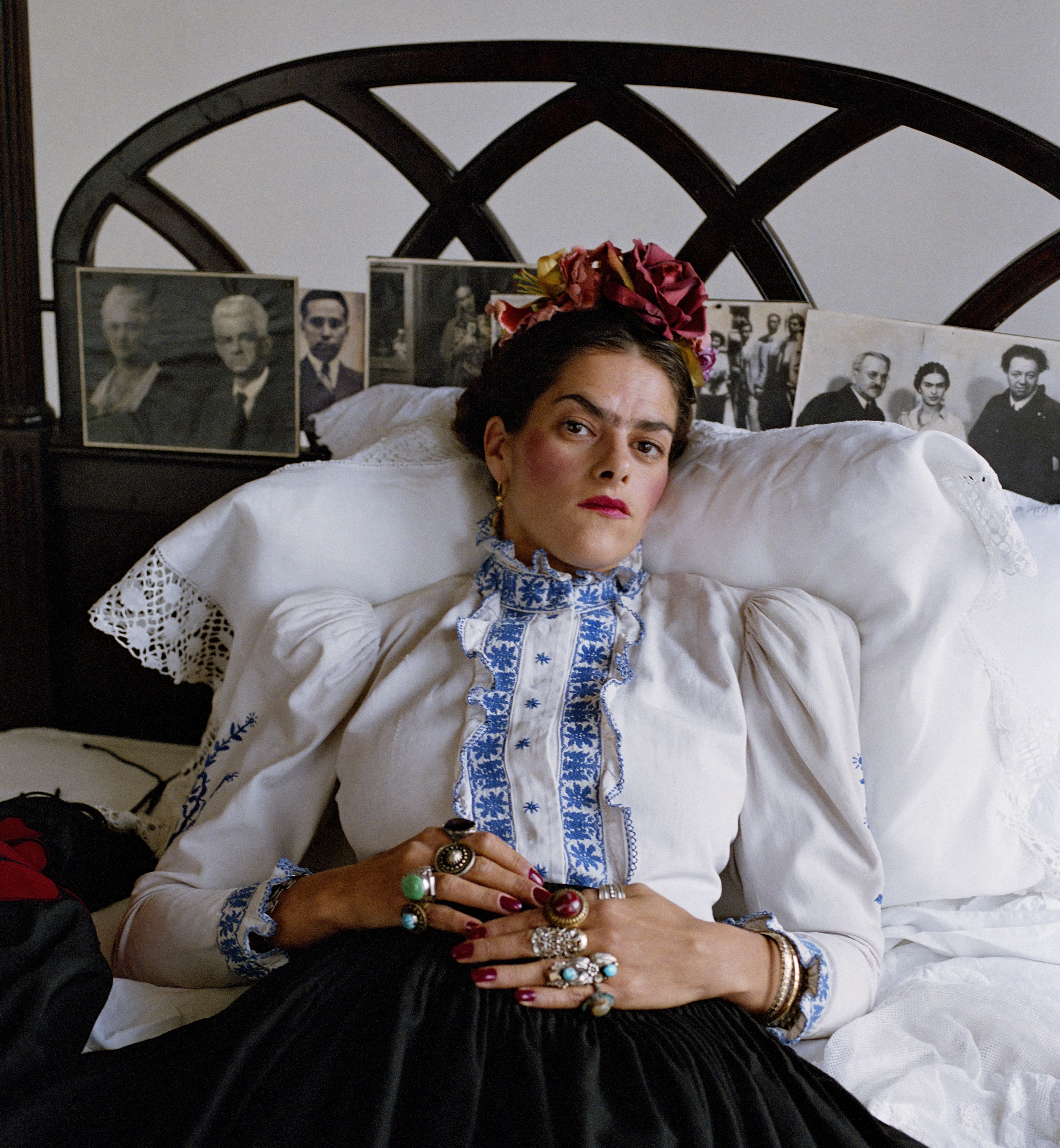 Tracey Emin dressed as Frida Kahlo, lying in a bed with several framed photos on the headboard.
