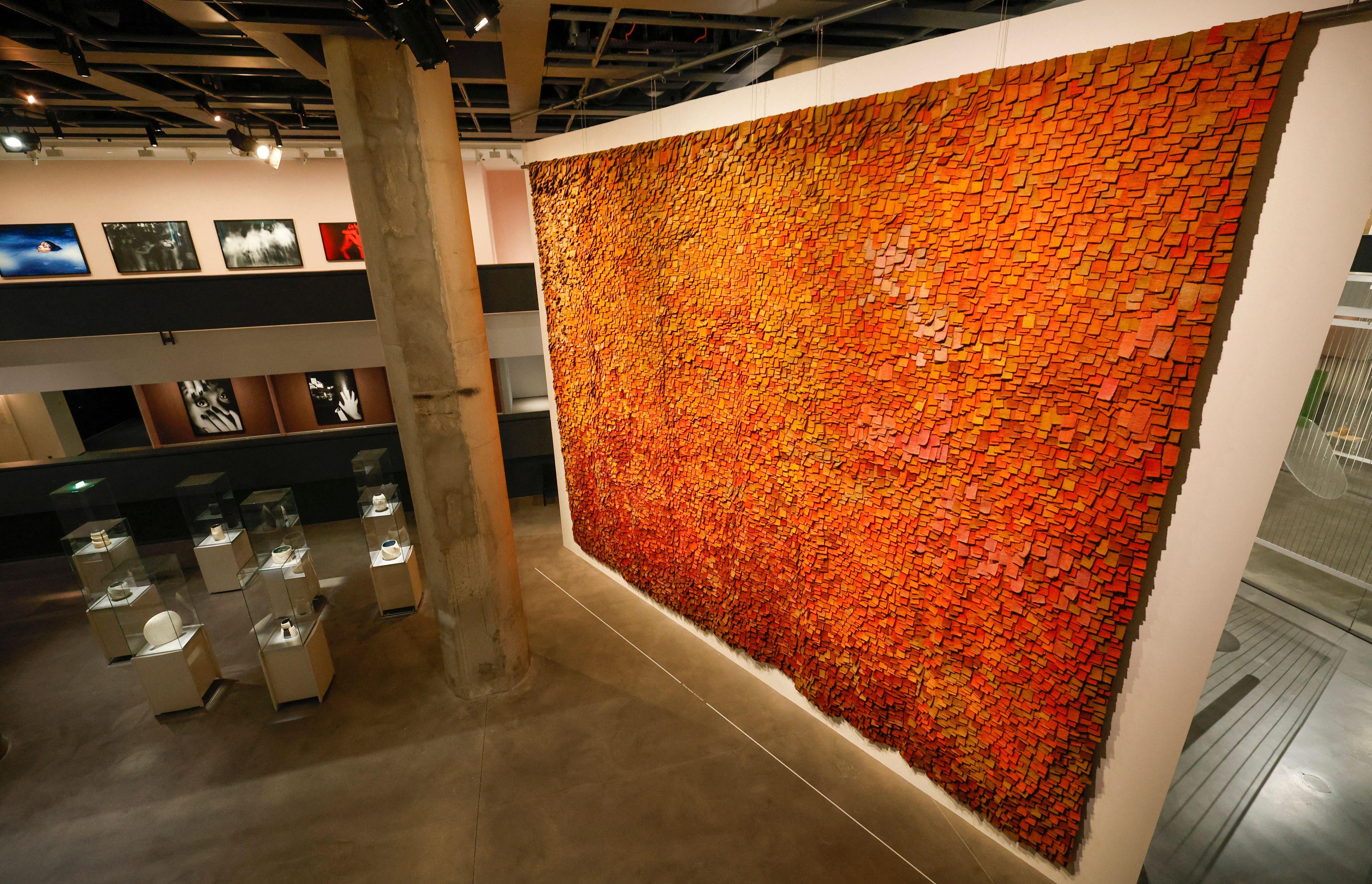 A large art installation made of orange and yellow square pieces hanging on a white wall in a museum.