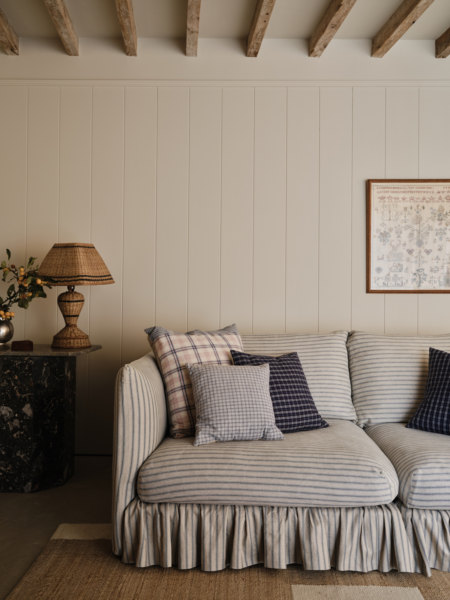 A cream-paneled room with exposed ceiling beams, featuring a striped sofa with patterned throw pillows, a wicker lamp on a marble side table, and framed embroidery on the wall.