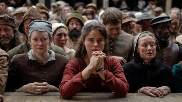 A still from the film Hamnet shows Jessie Buckley in the middle of an audience in Tudor dress. Her hands are clasped together and she is wearing a red dress