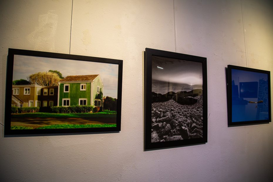Three framed photographs hang on a white wall: a green house, a black-and-white rocky landscape, and a blue abstract image.