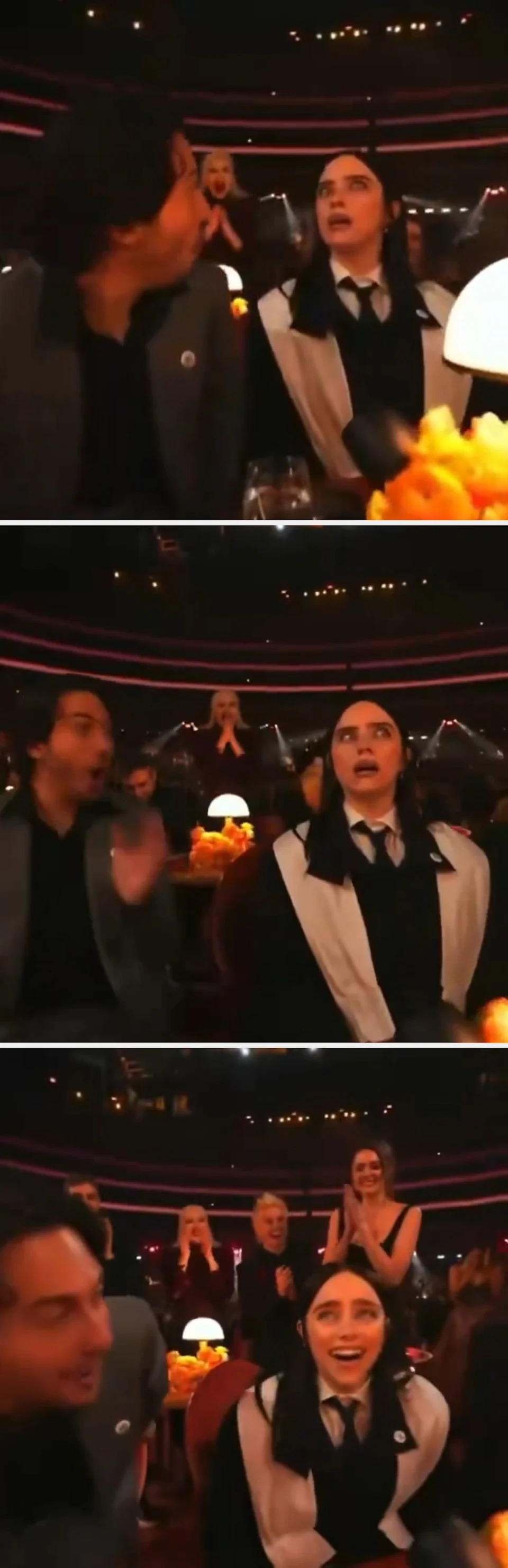 People seated at an event table, one wearing a suit and the other a black and white outfit, looking surprised. Warm lighting and an applauding crowd