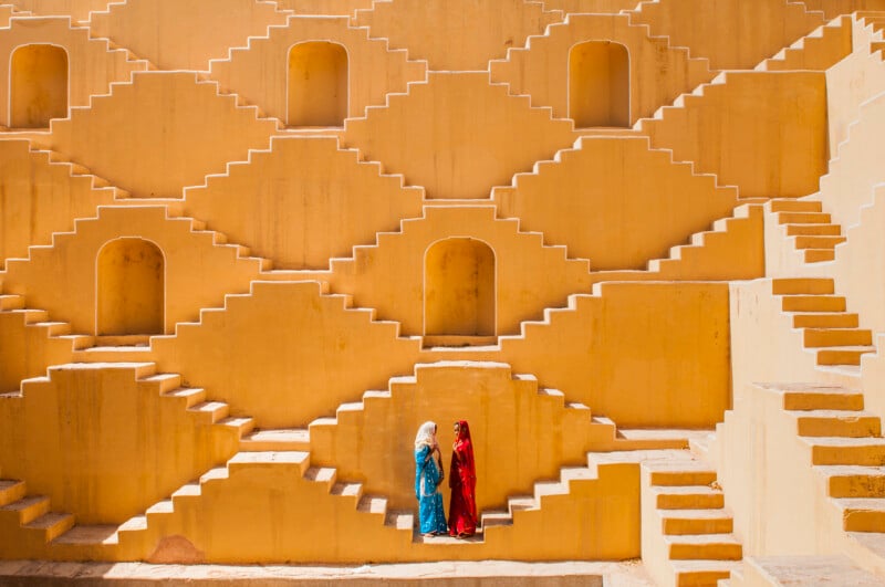 Two women in colorful traditional clothing stand on yellow geometric steps of an architectural structure with repeating patterns and doorways, creating a striking visual contrast.