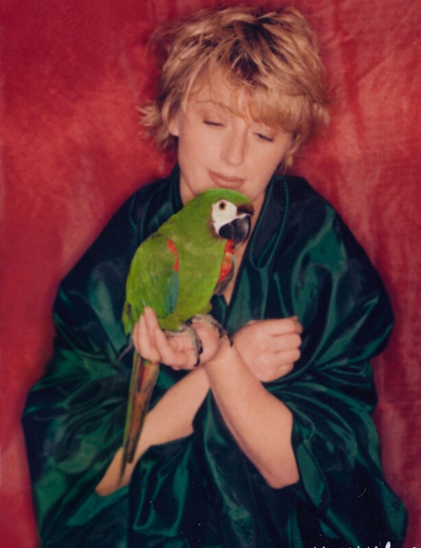 A person with short blond hair, wearing a green robe, gently cradles a green parrot with red and blue markings against a red background.