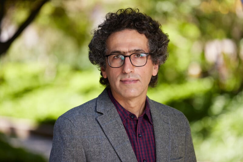 A man with curly dark hair, glasses, and a gray blazer over a red checkered shirt is standing outdoors with greenery and trees blurred in the background.