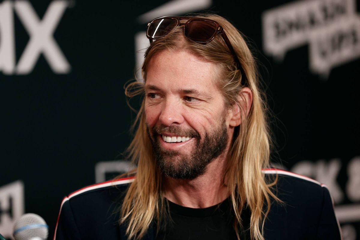 An individual with long blonde hair and a beard, wearing a black jacket, is seated and smiling in front of a microphone. The backdrop features a dark surface with various textual elements.