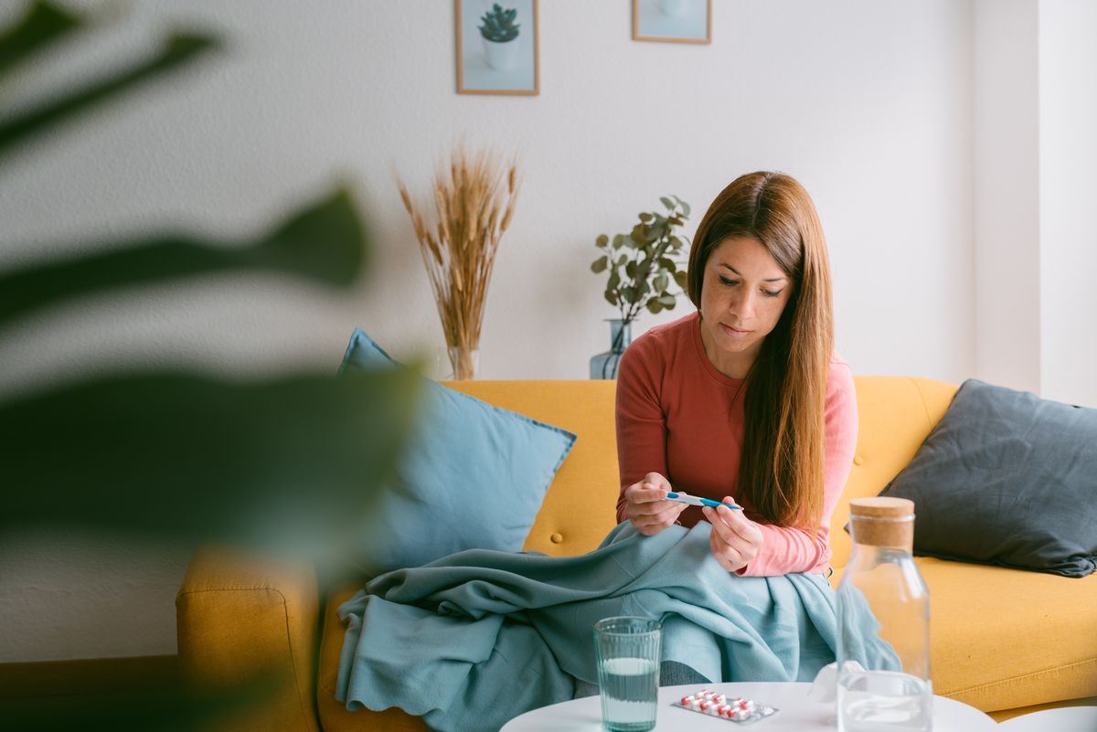 Sick young Caucasian woman sitting on the sofa at home wrapped in a blanket as she goes to take a pill to recover
