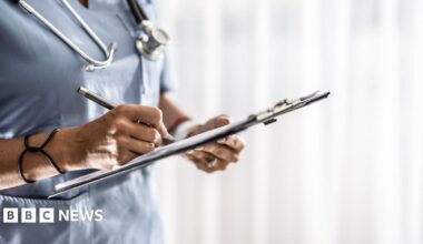 A woman in light blue nurses scrubs has a stethoscope around her neck. She is writing on paper on a clipboard.
