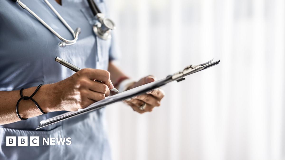 A woman in light blue nurses scrubs has a stethoscope around her neck. She is writing on paper on a clipboard.