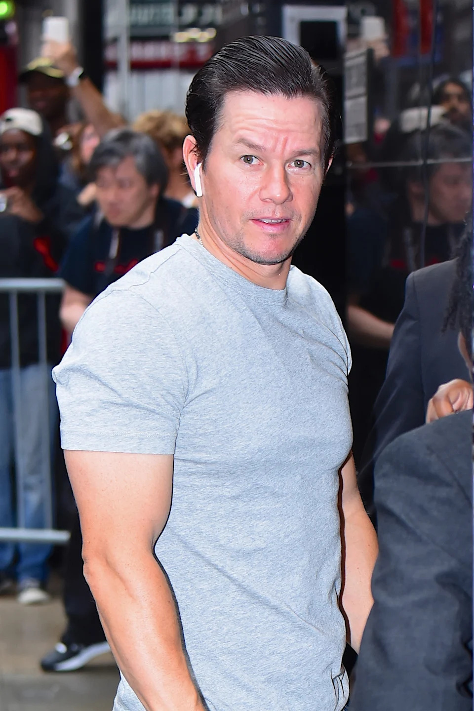 Mark Wahlberg in a casual gray t-shirt is walking outside, wearing earbuds, with people and barriers in the background