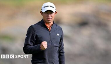 Collin Morikawa clenches his right fist after sinking his winning putt. He is wearing a white cap and black zipped-up top.
