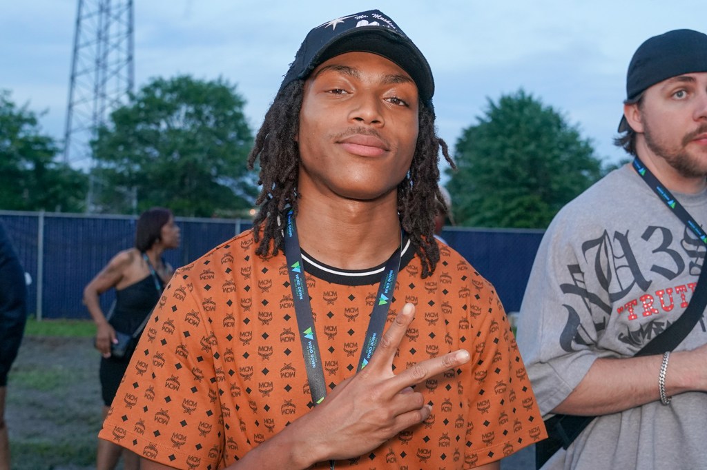 A young man wearing a black cap and an orange MCM t-shirt makes a peace sign to the camera.