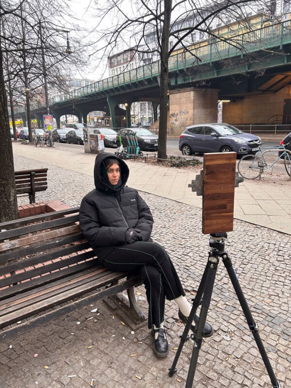 A person in a black winter jacket sits on a bench outdoors, facing a vintage box camera on a tripod. The street scene includes parked cars, bicycles, bare trees, and an elevated railway in the background.