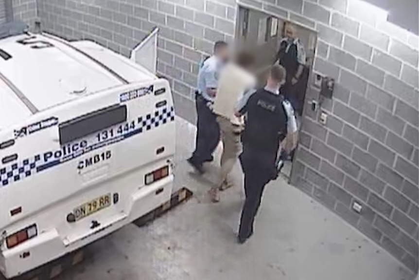 A man whose features are blurred exists a police car and is led by officers into the lockup.