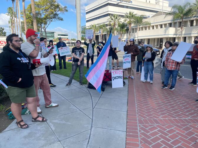 Speakers holding signs and flags supporting health care for trans youth gather around a man speaking into a microphone.