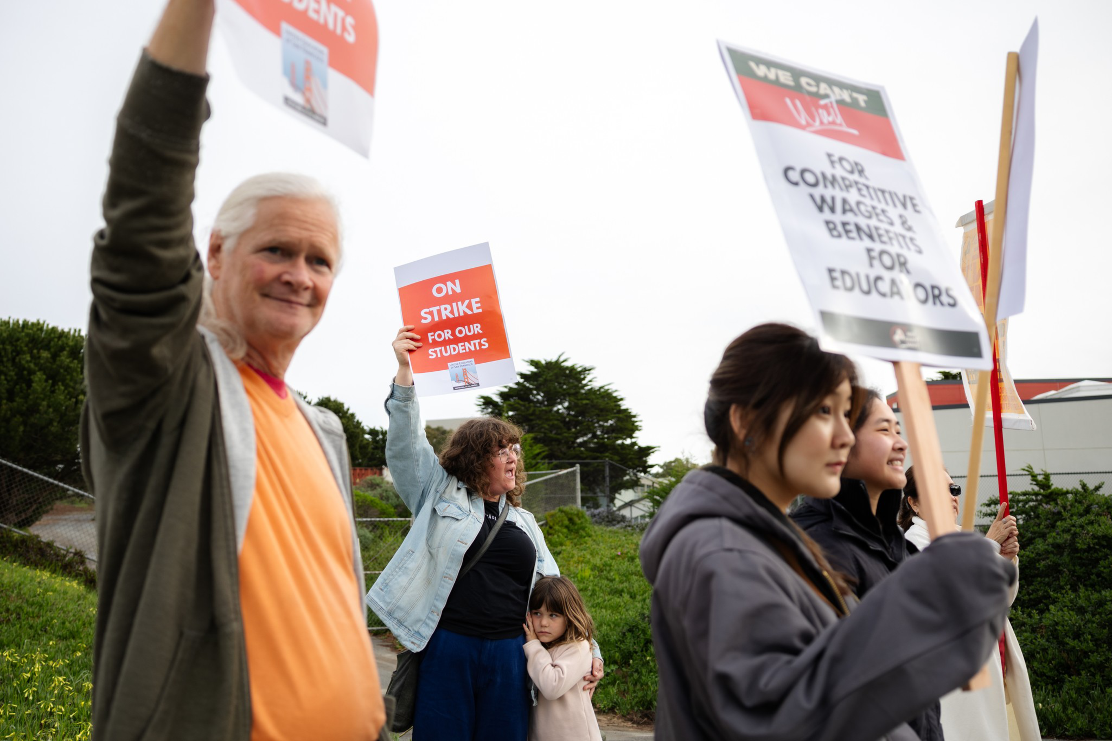 People of various ages hold signs advocating for better wages and benefits for educators while standing outdoors, with one woman hugging a young child.