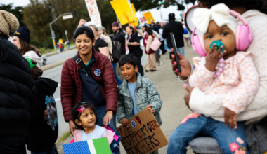 A diverse group of people, including children holding signs, gather outdoors in support of teachers during a peaceful protest or rally.