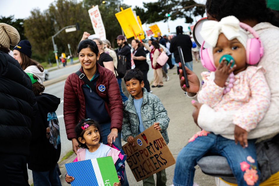 A diverse group of people, including children holding signs, gather outdoors in support of teachers during a peaceful protest or rally.