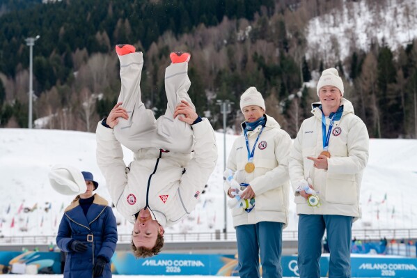 Silver medalist Ben Ogden, of the United States, does a back flip from the podium while gold medalist Johannes Hoesflot Klaebo, of Norway, and bronze medalist Oskar Opstad Vike, also of Norway, right, look on after the cross-country skiing men's sprint classic at the 2026 Winter Olympics, in Tesero, Italy, Tuesday, Feb. 10, 2026. (AP Photo/Evgeniy Maloletka)