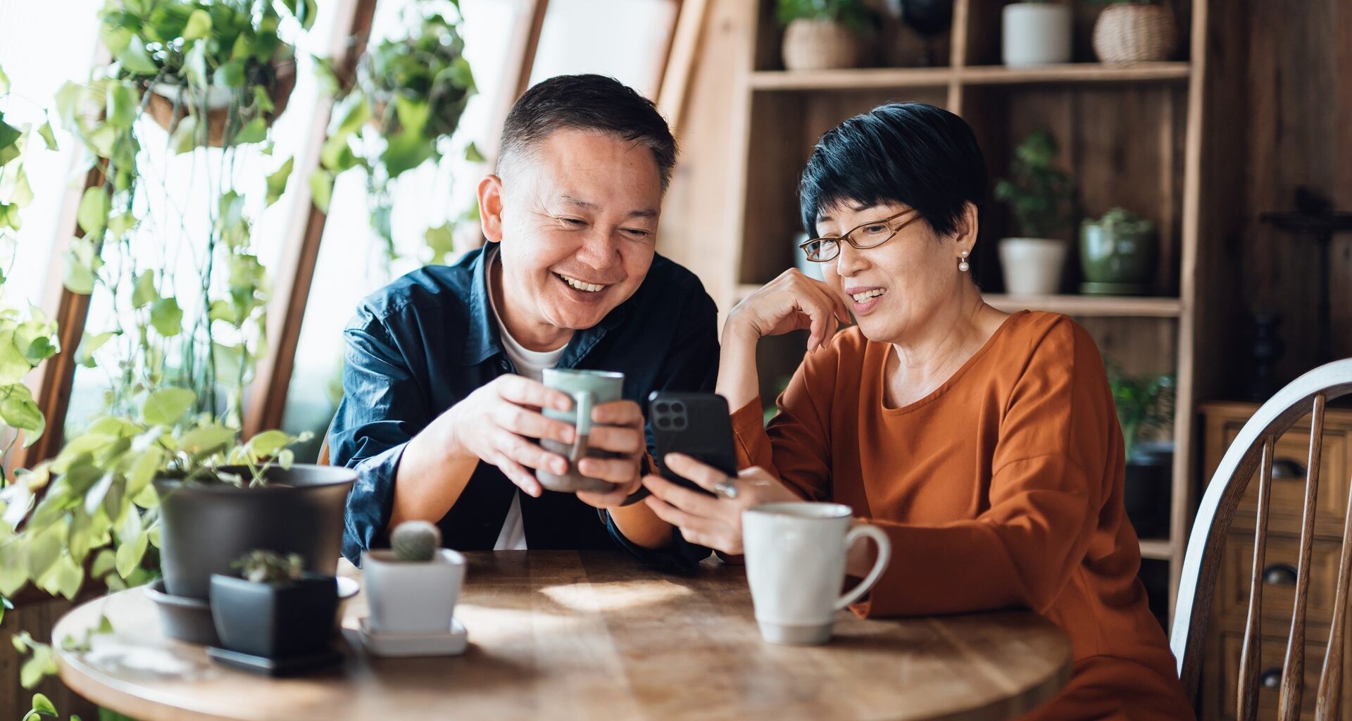 A happy retired couple smiles and drinks coffee while sitting at their kitchen table with plants next to them