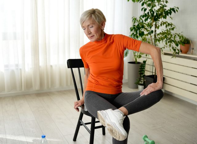 An elderly woman in an orange shirt and gray leggings sits on a chair, performing a seated stretch. She crosses her leg and twists her torso in a bright living room, promoting fitness and flexibility.