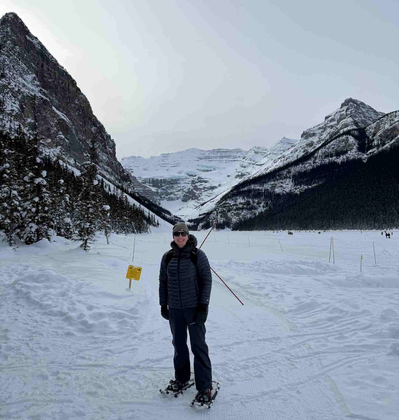 A person wearing winter gear and snowshoes stands on snowy ground with mountains in the background.