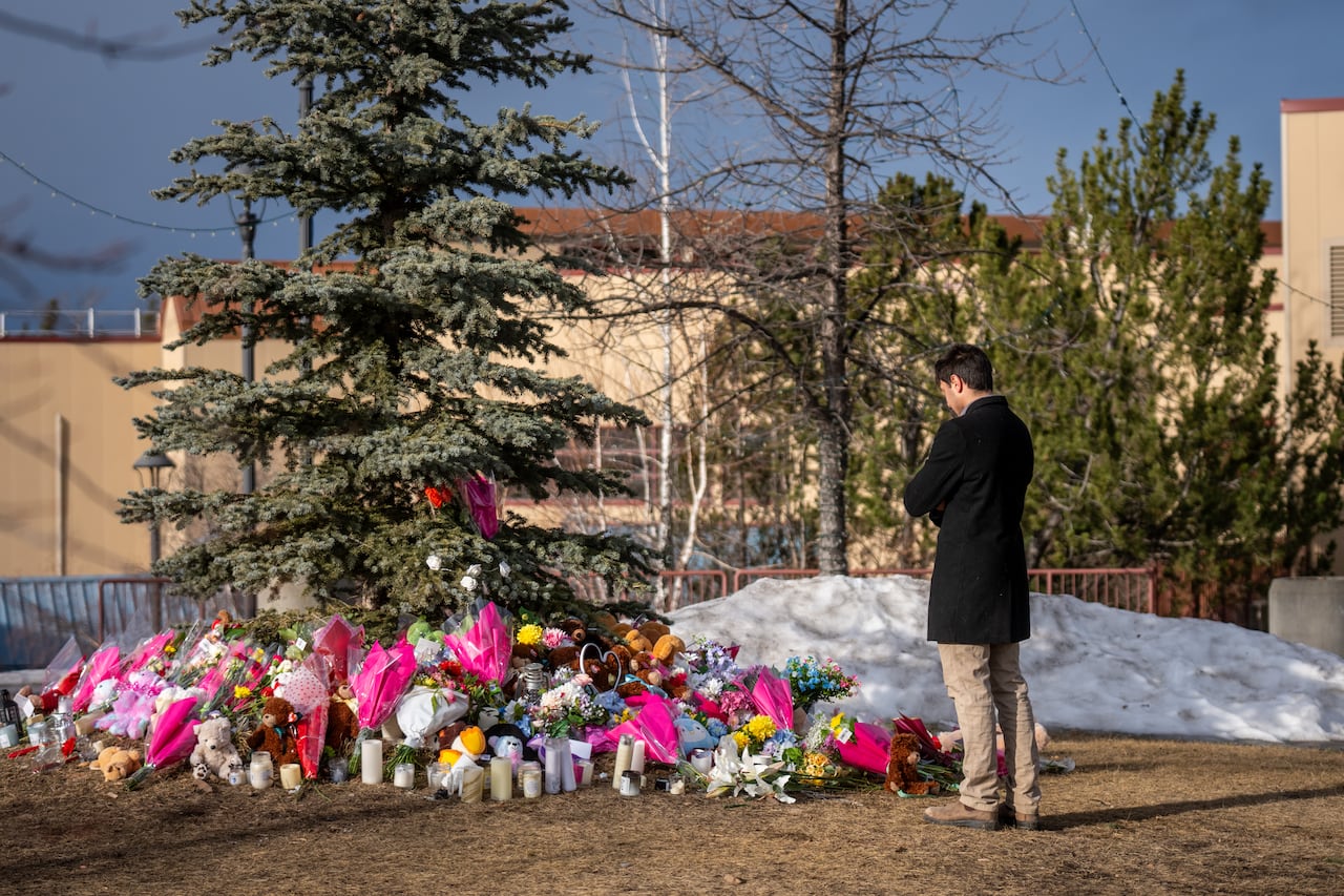 A man with his head bowed at a makeshift memorial with bouquets of flowers on the ground around a tree.