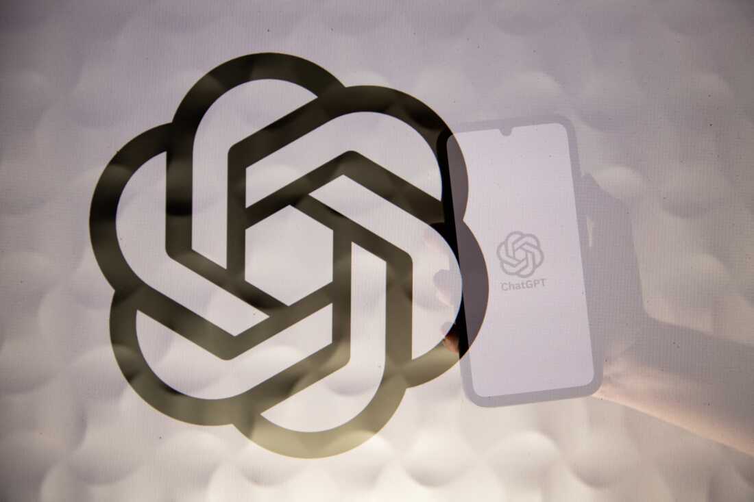 Double exposure photograph of a person holding a telephone displaying the ChatGPT artificial intelligence logo and the OpenAI company logo in front of a white lit background.