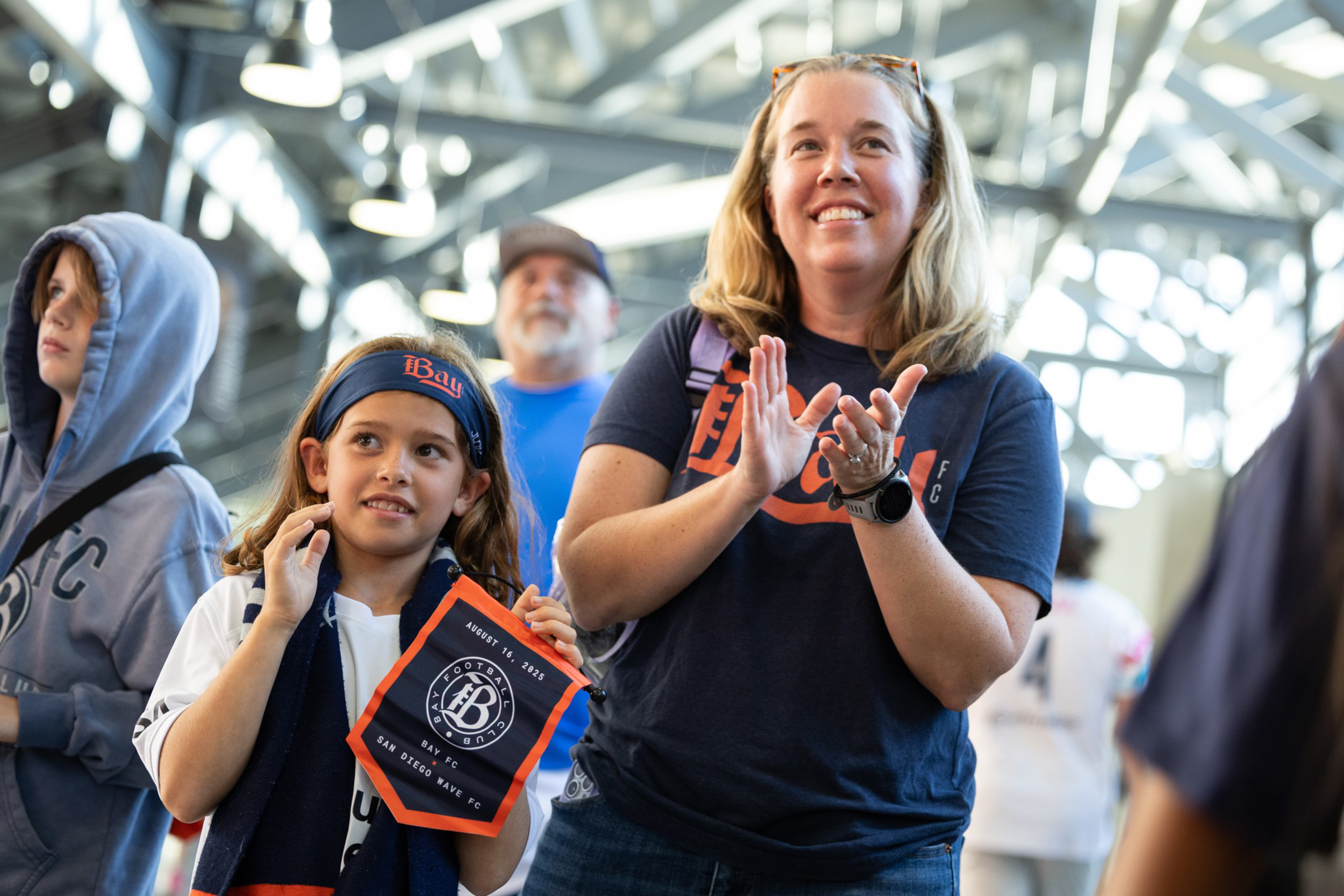 A smiling woman claps while a young girl beside her holds a Bay FC pennant at a bright indoor event with other attendees in the background.
