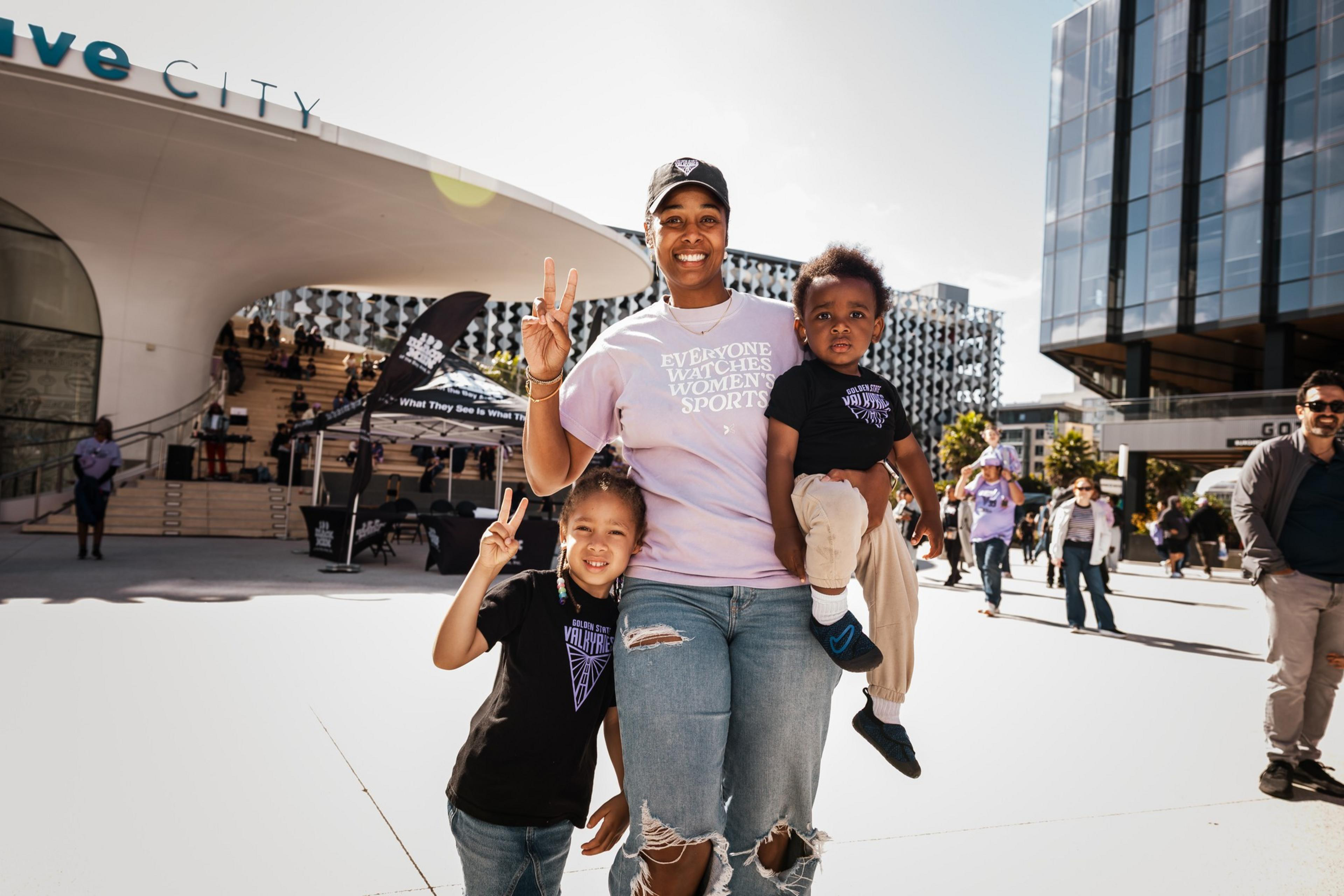 A woman smiles and holds a child while standing next to another child, all making peace signs outside a modern building on a sunny day.