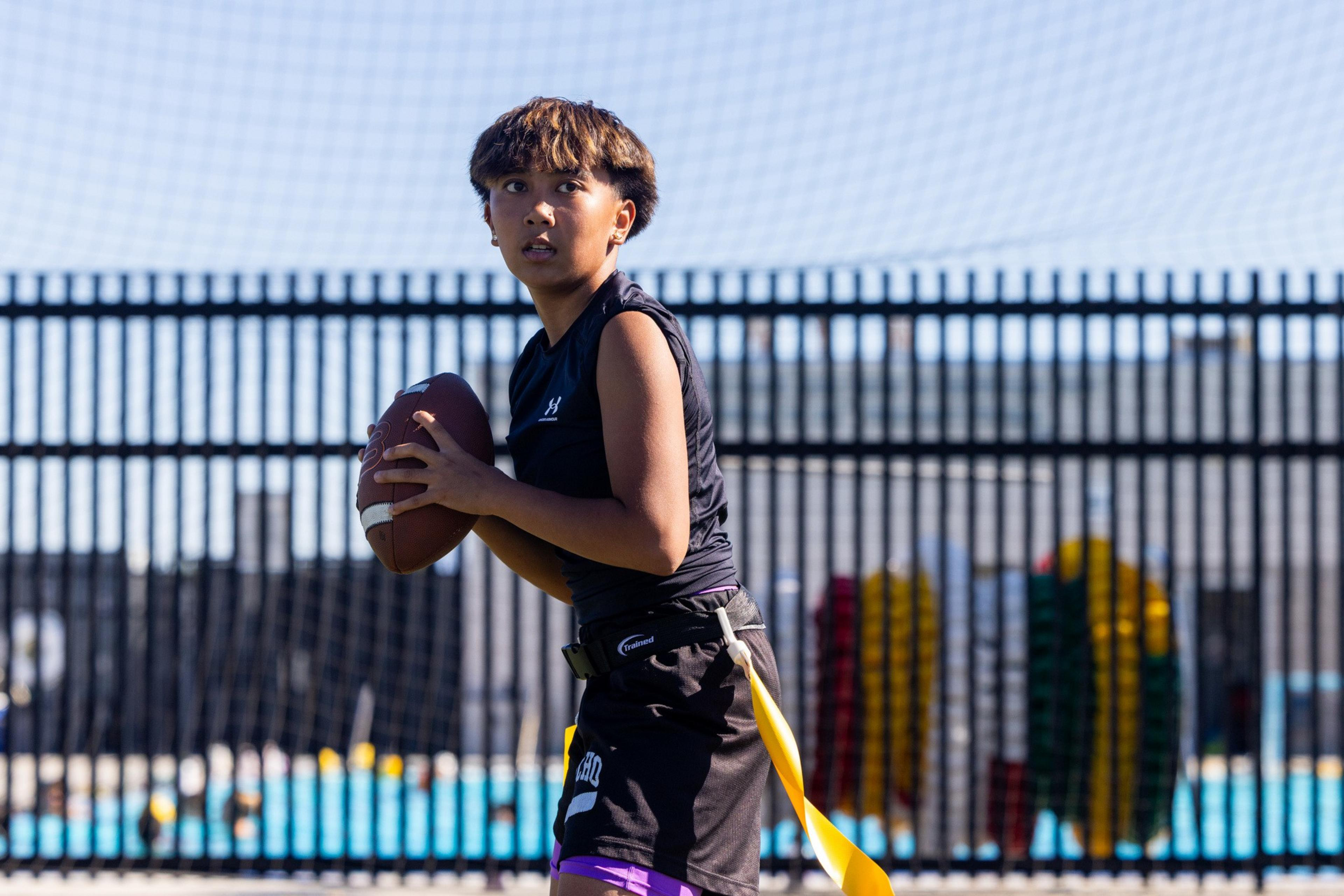 A young person in black athletic gear holds a football, preparing to throw, with a yellow flag hanging from their waist on a sunny field.