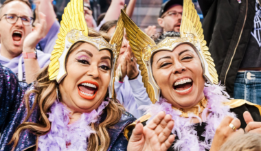 Two women wearing golden winged helmets and purple feather boas laugh and cheer enthusiastically in a crowded, lively setting.
