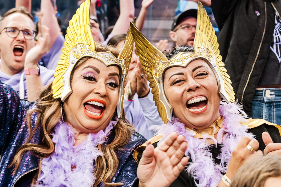 Two women wearing golden winged helmets and purple feather boas laugh and cheer enthusiastically in a crowded, lively setting.