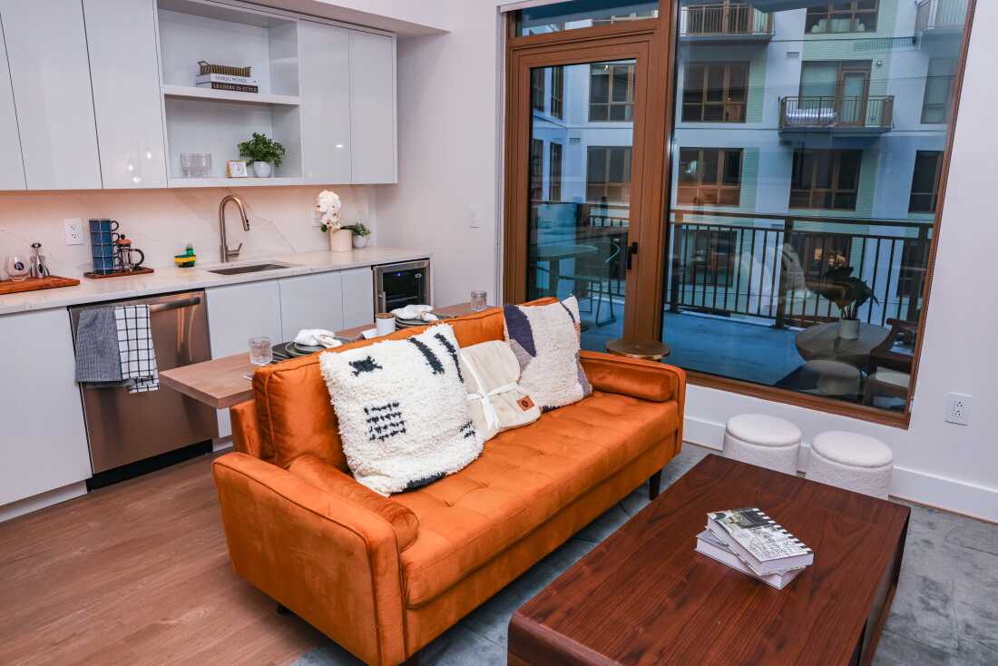 An orange couch and coffee table are set up near a small kitchen with a view of glass doors that open to a small patio overlooking another high-end residential building.