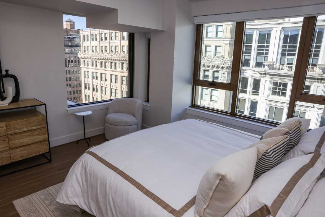 Two large windows in the bedroom of an Accolade penthouse model apartment overlook office buildings.