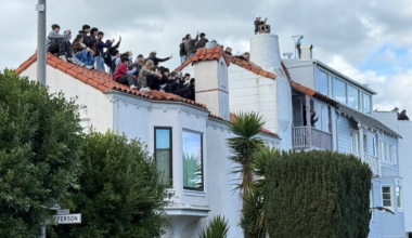 A large group of people sit closely together on a red-tiled rooftop, many holding up phones to capture photos or videos.