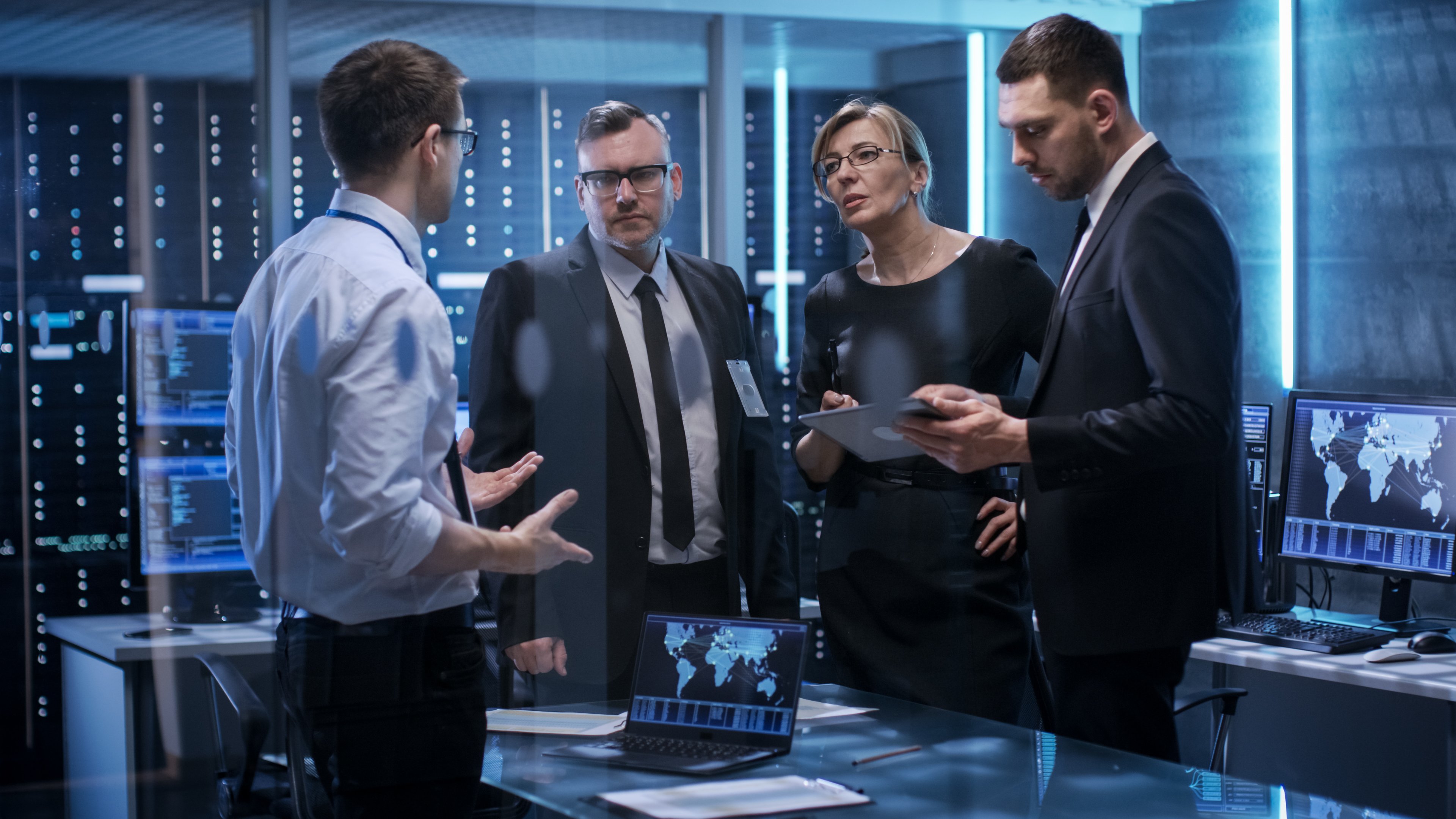People speaking in dimly-lit room with servers in the background.