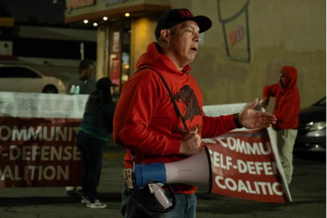 A man with medium skin tone, wearing a red hoodie with a design on it, speaks while holding a megaphone with a strap over his shoulder. There are people behind him holding up red banners.