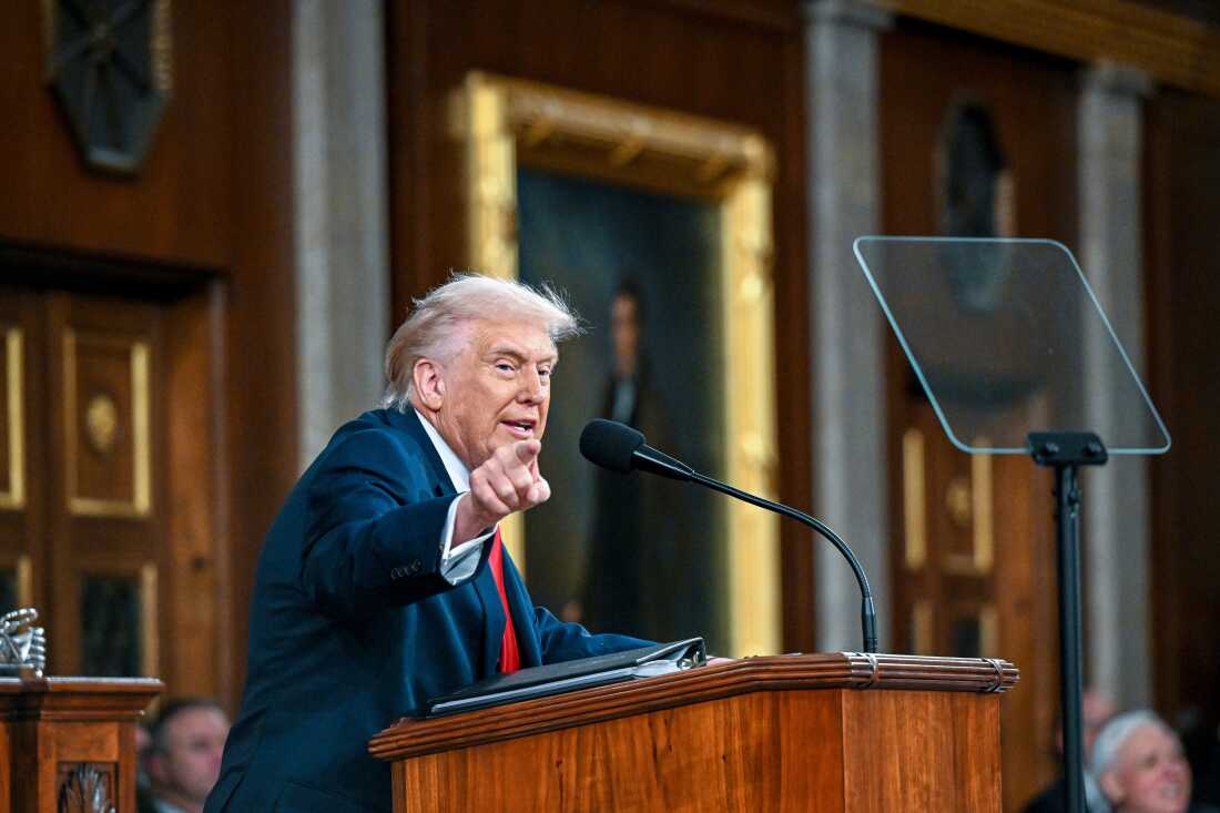 President Trump is pictured in a midrange photo at a lectern. A microphone and teleprompter are visible; he is pointing.