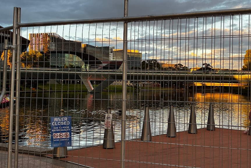 Wire fences and a small sign attached to a bollard on a riverbank, with the River bridge in the distance