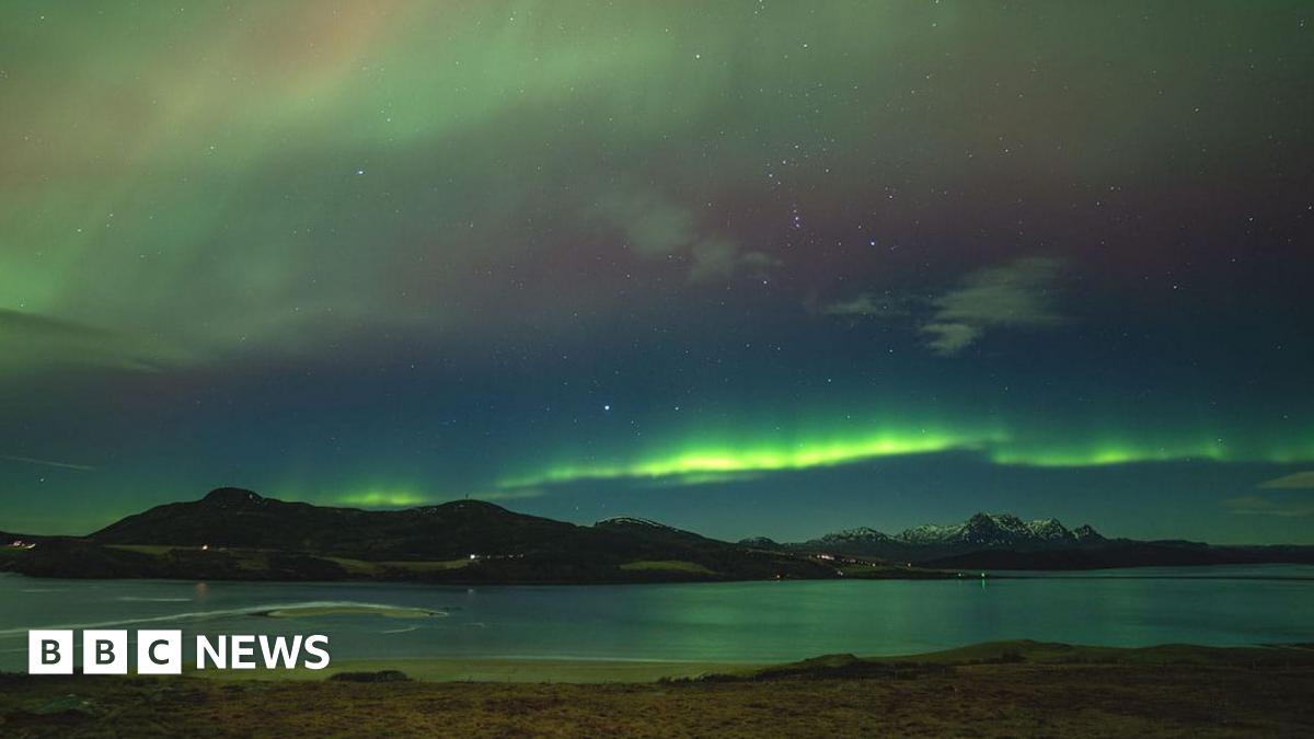 The Northern Lights in the north coast of Scotland. There are green and red stripes across the sky with snow-capped mountains below.