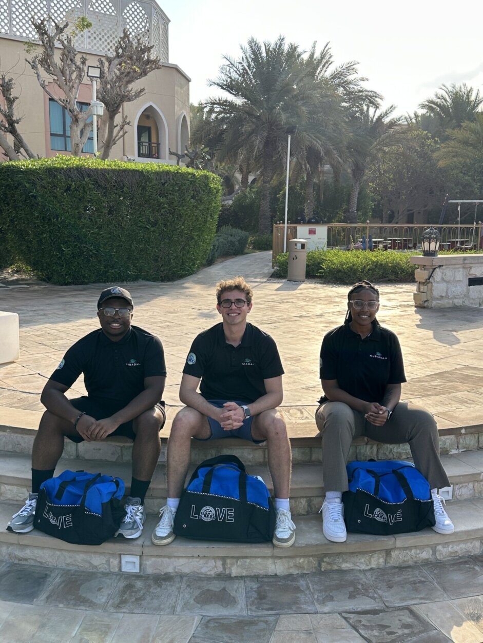 three young people sitting on a curb with duffel bags between legs.