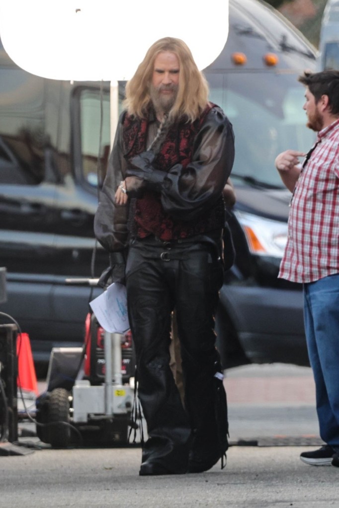 Will Ferrell in costume as a character with long blonde hair and a beard, wearing a black sheer shirt, a red and black vest, and black leather pants, on a film set.