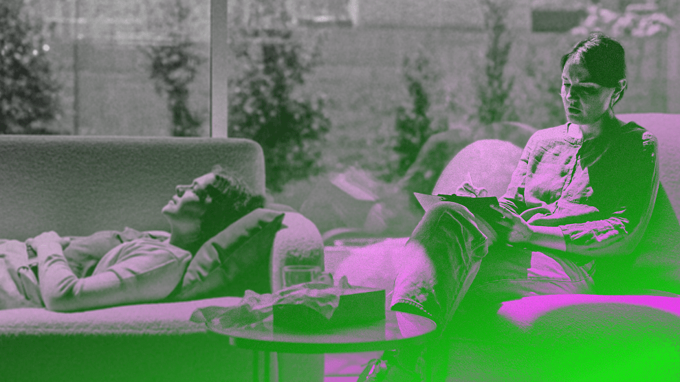 A photo of a psychologist taking notes as a patient lies on a couch. Both are Caucasian women with dark hair in a well-lit room.