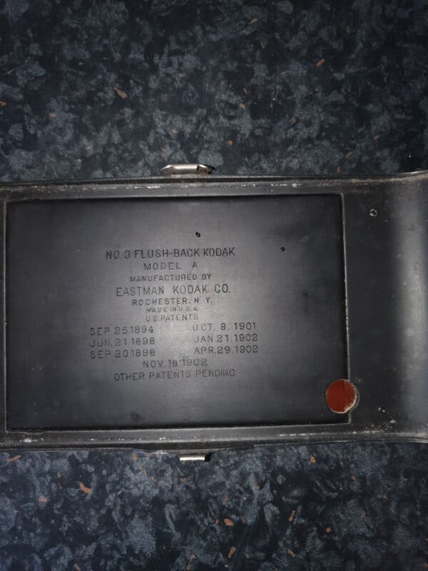 A metal plate inside a vintage camera reads: "No. 3 Flush-Back Kodak Model A, manufactured by Eastman Kodak Co., Rochester, N.Y., U.S.A." with several patent dates listed below. The camera is open, showing the plate.