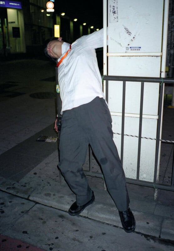 A man in office attire stands awkwardly slumped against a pole at night on a city sidewalk, his head tilted back and face partially obscured, suggesting exhaustion or sleep.